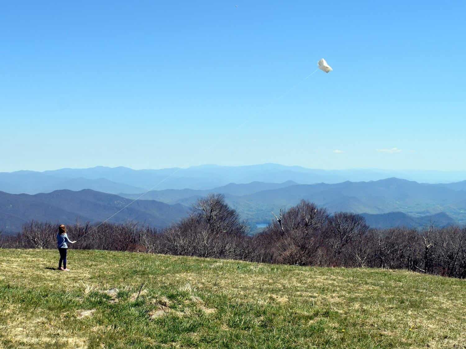 Girl flying kite in a grassy field bald on top of mountain with rolling mountains stretching in the distance